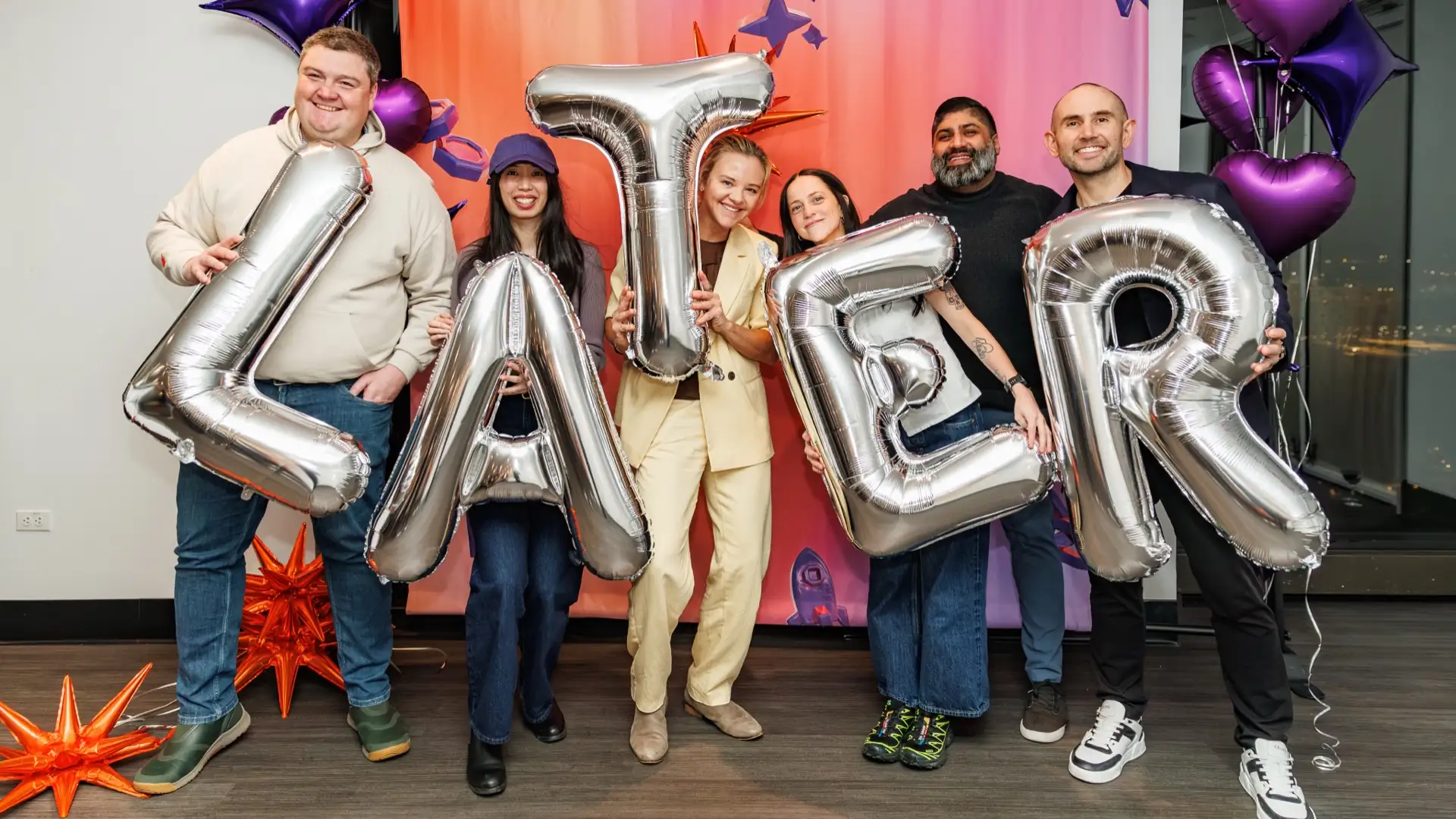 Six Later teammates smiling and holding giant silver balloon letters that spell LATER, in front of a pink and orange backdrop with purple balloons.